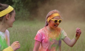 a girl covered in coloured powder wearing yellow sunglasses and headband laughs happily