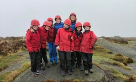 A group of boys and girls in hard hats and overalls stand at the top of a rocky outcrop in the Peak District